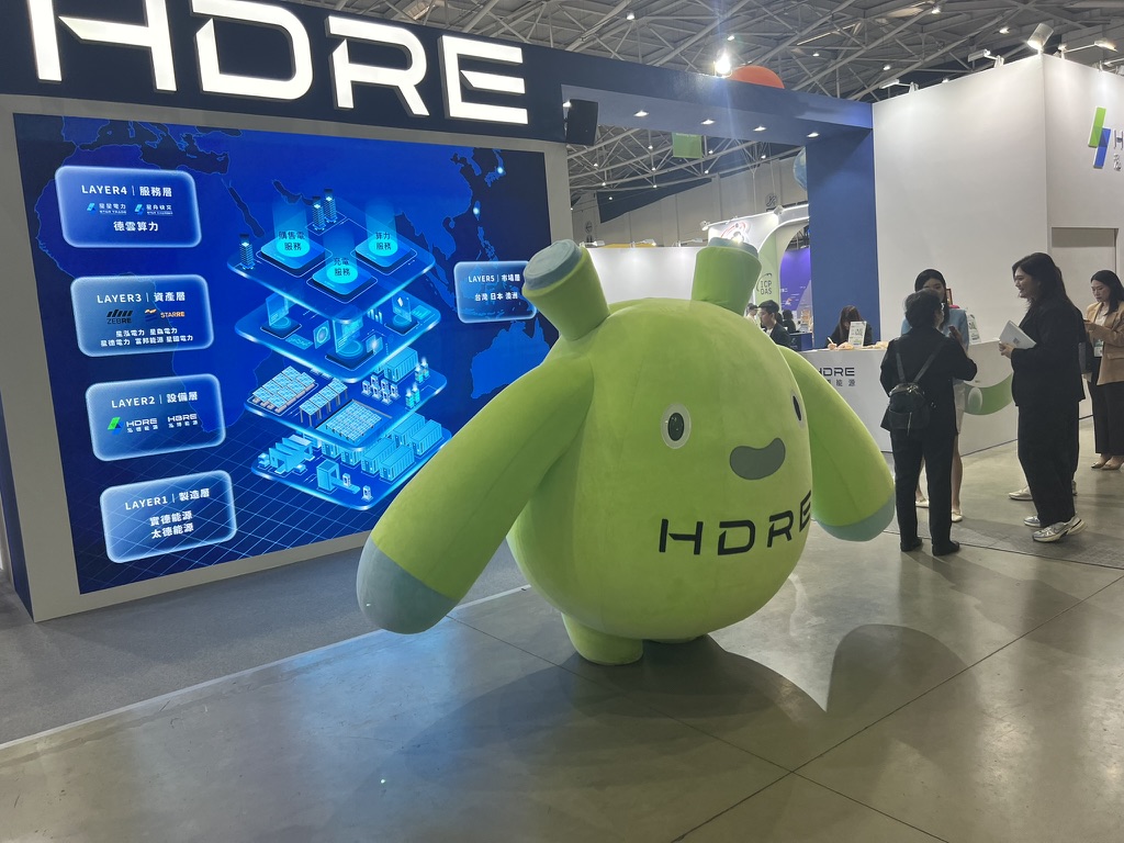 A green mascot in front of a power-systems display on the Energy Taiwan exhibition floor.